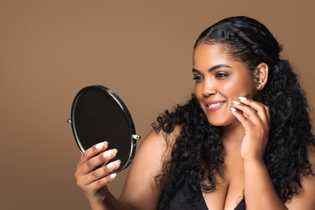 Beautiful latin plus size woman looking at small mirror, enjoying her brand new organic skin care routine, standing over brown studio background.